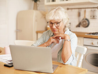 Modern technology, old people and retirement. Gray haired female pensioner in glasses filling in loan application form online, sitting in front of laptop, reading information with serious focused look